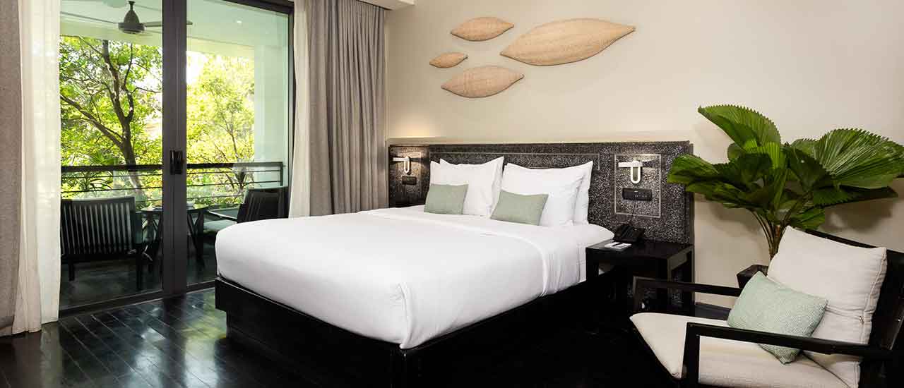 Modern hotel room with a king-sized bed, white linens, black headboard, decorative wall art, and a balcony overlooking greenery.